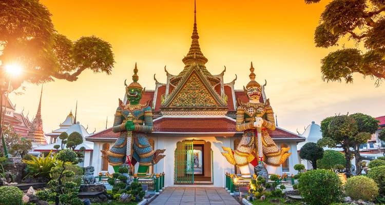 Wächterfiguren bei Sonnenuntergang vor einem lebhaft gefärbten thailändischen Tempel mit orangefarbenem Himmel