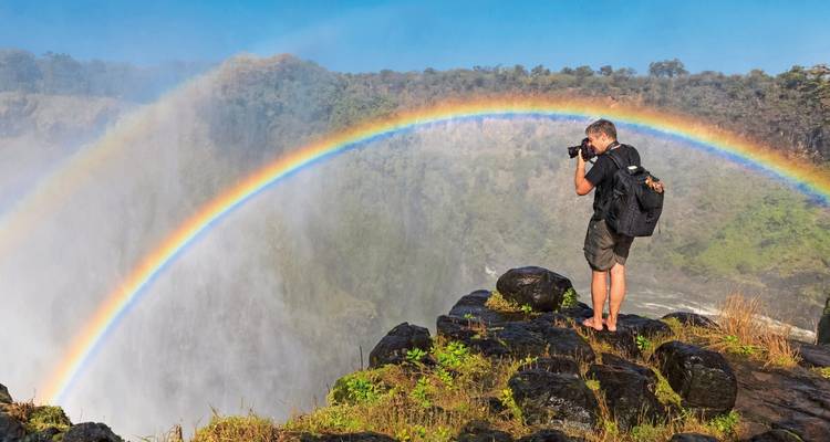 Un photographe capture un arc-en-ciel au-dessus des chutes Victoria depuis le bord d'une falaise rocheuse dans la brume des embruns.