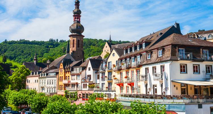 Des maisons à colombages colorées bordent une rue riverside dans une vallée allemande verdoyante, dominées par la haute tour d'une église baroque.