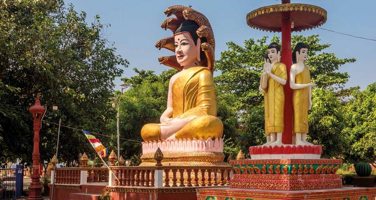 Sanctuaire bouddhiste extérieur coloré présentant une grande statue de Bouddha doré assis entouré de décorations ornées et d'arbres verts luxuriants sous un ciel bleu éclatant.