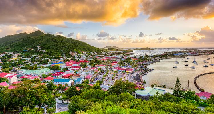 Vue panoramique du coucher de soleil sur une ville côtière caribéenne colorée avec collines, port et yachts ancrés.