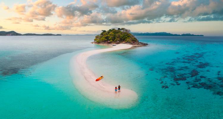 Un minuscule banc de sable idyllique s'incurve dans une eau turquoise vers un îlot luxuriant tandis qu'un couple et un kayak reposent sur le sable blanc au coucher du soleil.