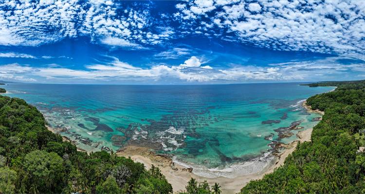 Vue aérienne panoramique d'une baie tropicale vierge aux eaux turquoise, aux récifs coralliens et à la côte de forêt tropicale luxuriante sous un ciel dramatique.