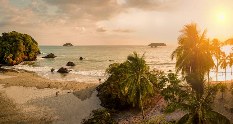 Baie tropicale panoramique avec des palmiers, de petites îles au large et des personnes se promenant sur le sable lors d'un coucher de soleil chaud.
