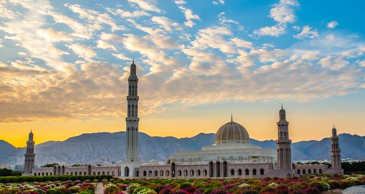 Sultan Qaboos Grote Moskee omgeven door kleurige tuinen met rotsachtige bergen bij zonsopgang.