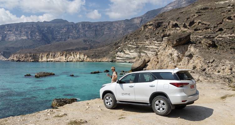 Een witte 4x4 geparkeerd op een rotsachtige kustklif met uitzicht op turquoise water en dramatische kalksteenheuvels in zuidelijk Oman.