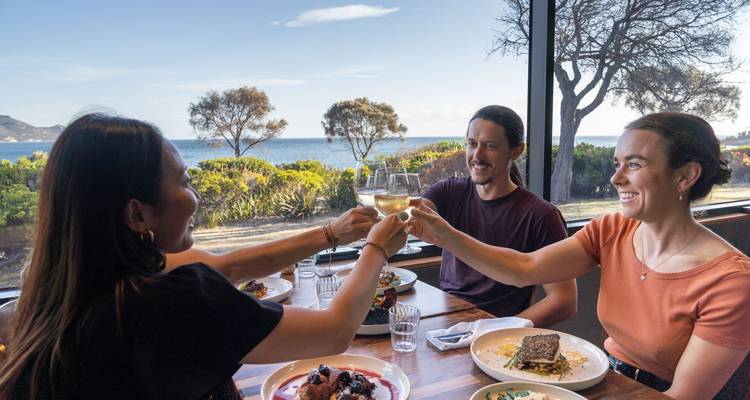 Trois amis portant un toast au vin blanc à une table près de la fenêtre donnant sur la mer et les arbres.