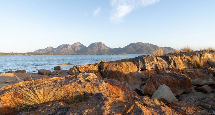 Des rochers couverts de lichen orange et baignés de soleil le long d'une baie calme avec une chaîne de montagnes lointaine.