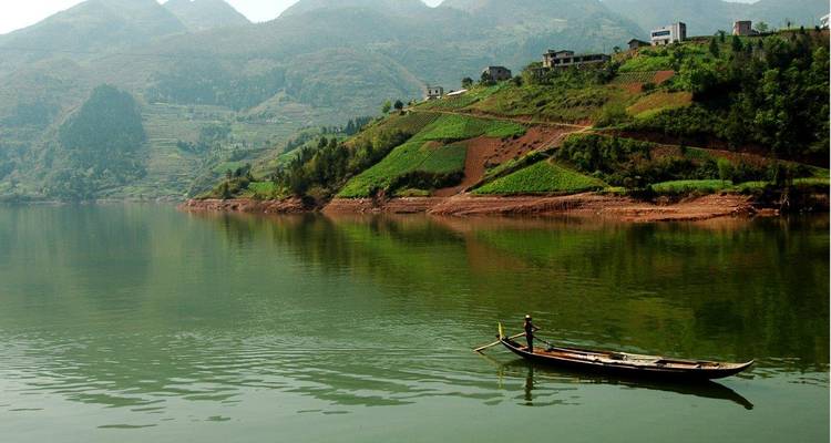 Small wooden boat with a lone rower glides across calm green water backed by terraced hills and misty mountains.