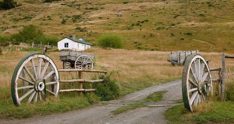 Rustikale patagonische Landschaft mit übergroßen hölzernen Wagenrädern, die eine Feldweg rahmen, der zu einem kleinen Bauernhaus führt.