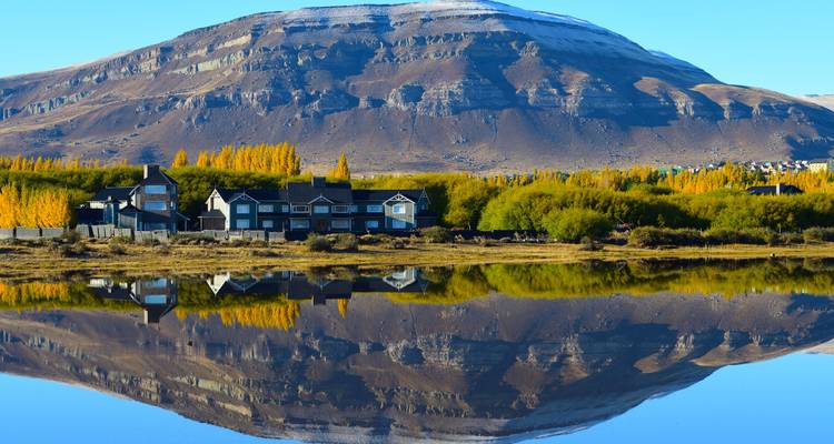 Ein ruhiger See spiegelt die Herbstbäume und einen zerklüfteten Berg hinter den Seeuferhaüsern perfekt wider.