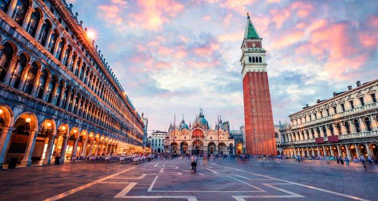 Der Markusplatz in Venedig in der Morgenröte mit dem Campanile-Turm und der Basilika, umrahmt von Arkadenbauwerken unter einem pastellfarbenem Himmel.