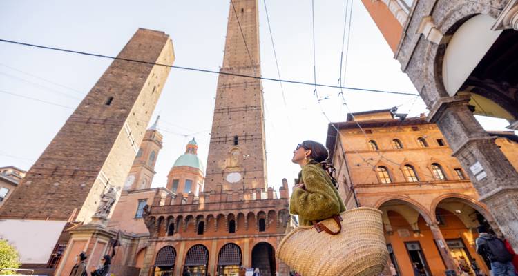 Vrouwelijke reiziger die omhoog kijkt naar Bologna's iconische Twee Torens tussen omringende historische gebouwen op een zonnige dag.