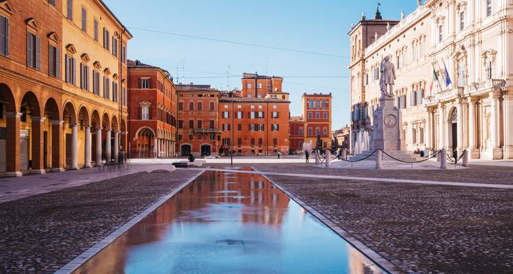Rustig plein in Modena met historische rode en okergele gebouwen weerspiegeld in een lang plas onder een heldere ochtendhemel.