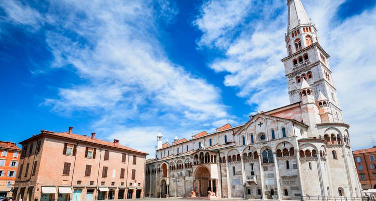 De Romaanse Modena Kathedraal en de hoge Ghirlandina klokkentoren tegen een levendige blauwe lucht en dramatische wolken.