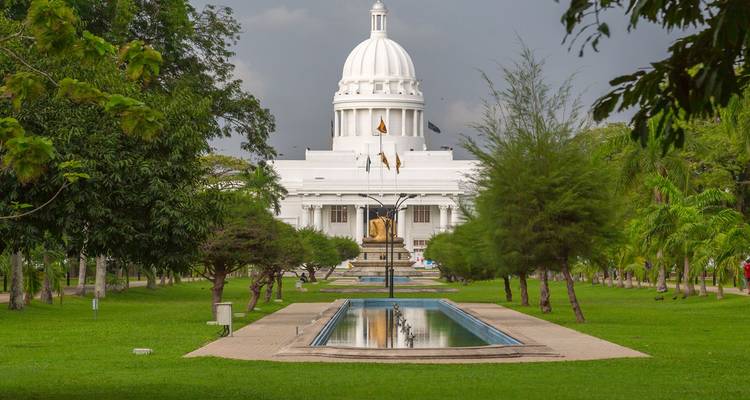 Blick entlang eines gepflegten Parkweges mit Spiegelbecken, der zum weißen, kuppelförmigen Colombo Town Hall Gebäude führt, das von Bäumen umrahmt wird.