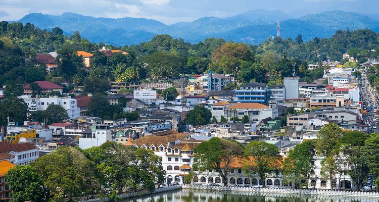 Panoramablick auf die Kandy-Stadtlandschaft mit Gebäuden, die sich um den See gruppieren, und bewaldeten Bergen im Hintergrund.