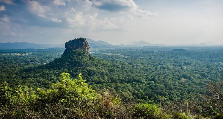Der ikonische Sigiriya Rock erhebt sich über einen dicht bewaldeten grünen Wald mit entfernten nebligen Bergkämmen unter einem dunstigem Himmel.