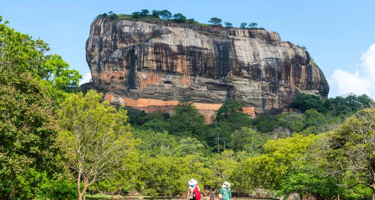 Die massive Felsfestung von Sigiriya erhebt sich über den Dschungel, während Touristen den Gartenweg entlanggehen.