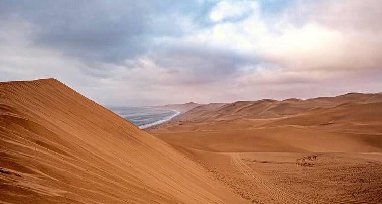 Sweeping orange sand dunes tumble toward the Atlantic coastline beneath a dramatic cloudy sky.