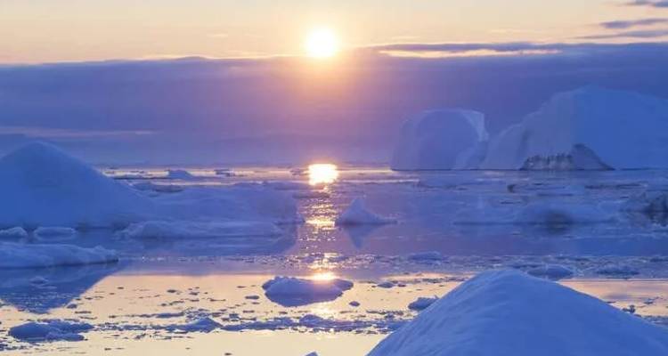 Arctische zonsondergang die weerspiegeld wordt op een rustige ijzige zee vol met blauwe ijsbergen en sneeuwheuvels.