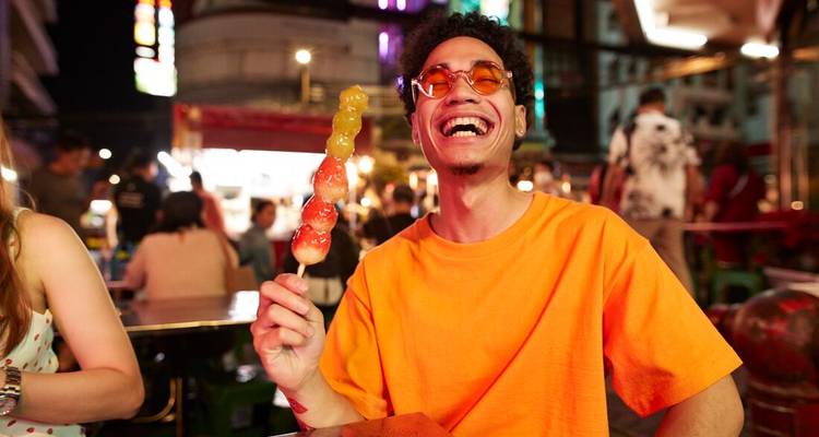 Young man in an orange shirt laughs while holding a colourful skewer at a lively Bangkok night market.