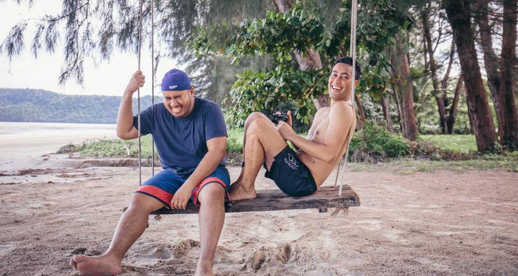 Two friends laugh while sitting on a wooden swing hanging from coastal trees above a sandy beach.