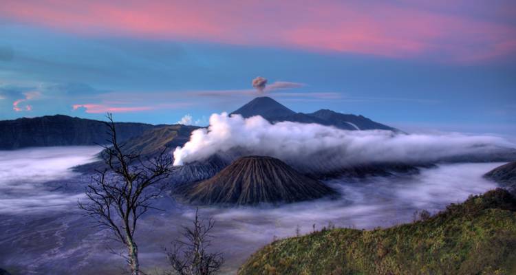 Impresionante paisaje volcánico del Monte Bromo emitiendo vapor blanco entre nubes arremolinadas y un cielo de amanecer lleno de colores.