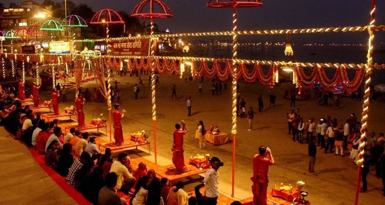 Abendliche Ganga-Aarti-Zeremonie auf den Varanasi-Ghats, beleuchtet durch Lichterketten in bunten Farben, mit sitzenden Menschenmengen.