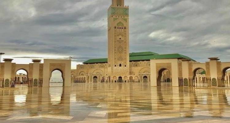 La Mosquée Hassan II à Casablanca se reflétant dans une cour en marbre trempée par la pluie sous un ciel chargé.