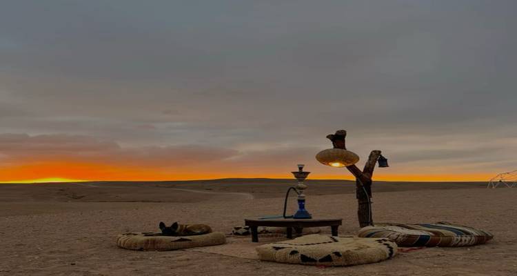 atardecer sobre una llanura desértica árida con cojines y una pipa de agua dispuestos alrededor de una mesita