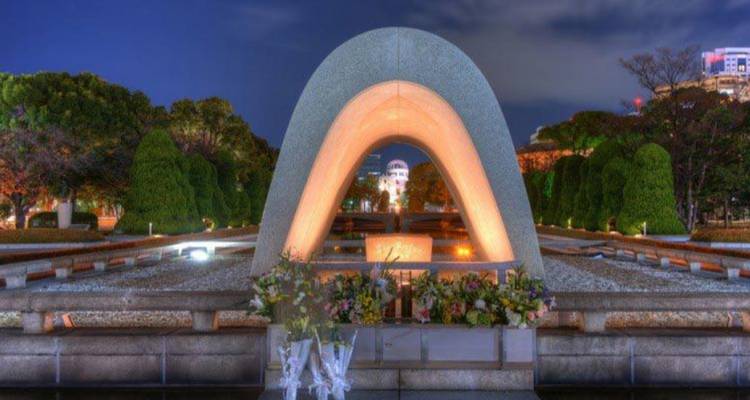 Beleuchteter Kenotaph-Bogen im Hiroshima-Friedenspark bei Nacht mit Gedenkdom dahinter
