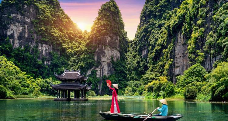 Ein traditionelles Boot gleitet durch dramatische Karstfelsen in Ninh Binh, während eine Frau in rotem Ao Dai mit einem Fächer winkt.
