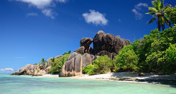 Sonniger tropischer Strand mit türkisfarbenem Wasser, weißem Sand und dramatischen Granitfelsen, umgeben von üppig grüner Vegetation.