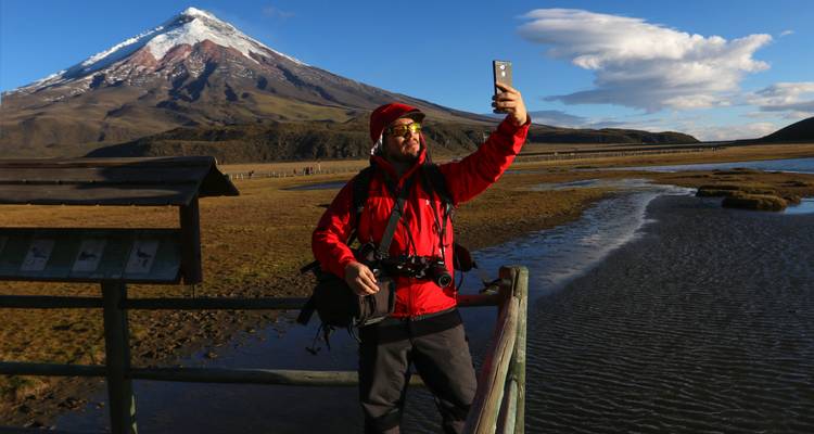 Reiziger in rood jasje maakt een selfie met de sneeuwbedekte vulkaan Cotopaxi die hoog achter hen uittorent.