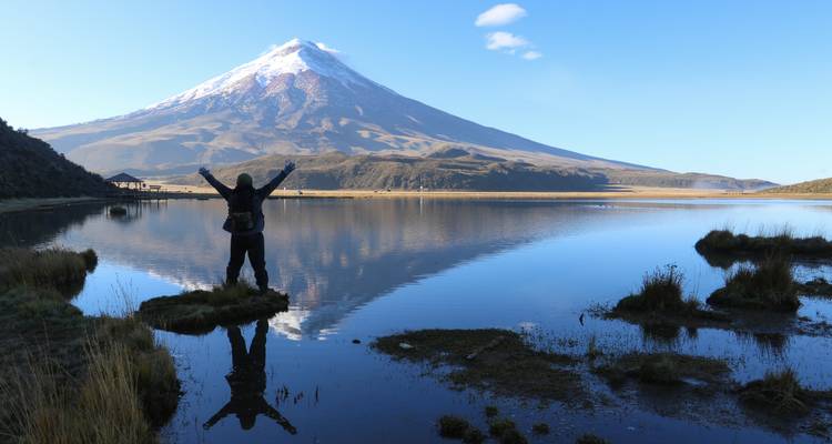 Een silhouet van een wandelaar met opgeheven armen naast een reflecterend meer met de vulkaan Cotopaxi gespiegeld in het water.