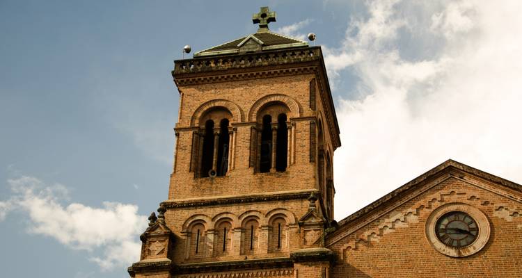 Bakstenen klokkentoren van een historische kerk met rond raam en kruis, tegen een gedeeltelijk bewolkte hemel.