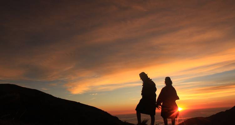 Paar in Silhouette, das sich an den Händen hält auf einem Grat und einen dramatischen Sonnenaufgang über den Anden beobachtet.