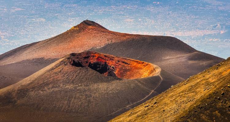 Kleurrijke vulkanische kratters en lavavelden op de hogere hellingen van de Etna met een stadslandschap dat vaag zichtbaar is ver beneden.