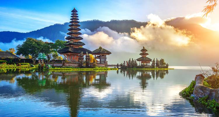 Le complexe du temple Ulun Danu Bratan au bord du lac se reflète dans des eaux calmes tandis que la lumière du lever du soleil traverse la brume matinale.