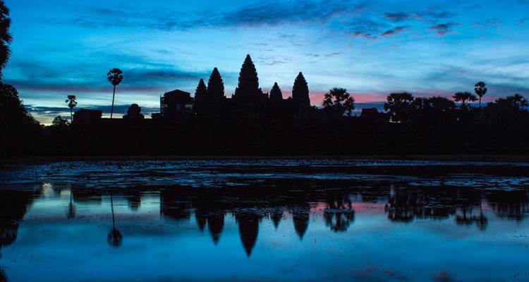 Silhouette of Angkor Wat towers reflected in a tranquil pond under a vibrant blue-pink sunrise sky.