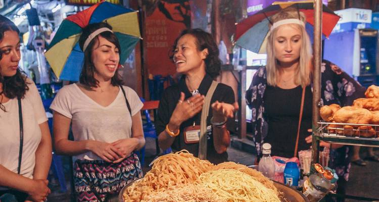 Tourists wearing colorful umbrella hats enjoy a lively street food stall filled with noodles and snacks.