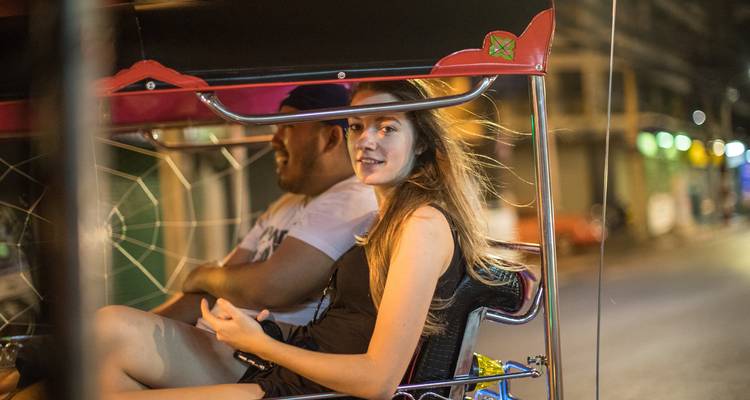 Une jeune femme voyage dans un tuk-tuk la nuit, les lumières de la ville floues derrière elle.