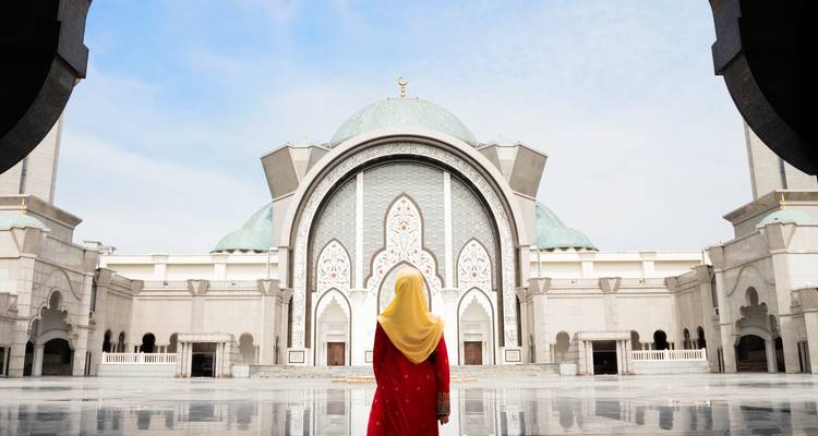 Une femme en robe rouge et hijab jaune se tient devant une vaste cour de mosquée aux dômes bleus imposants.