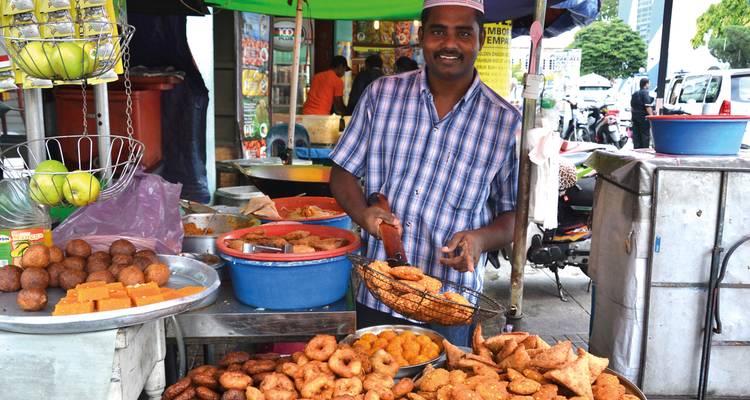Vendeur de restauration rapide souriante à un étal coloré présentant une variété de collations frites et de sucreries dans un marché ouvert.