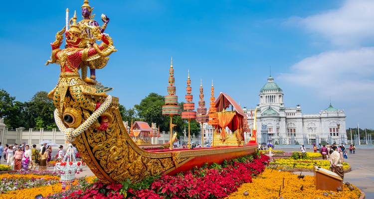 Sculpture élégamment dorée d'une barge royale entourée de fleurs colorées avec le Palais du Trône Ananta Samakhom en arrière-plan.