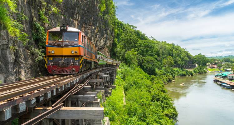 Un train aux couleurs vives circulant sur un viaduc en bois construit contre une falaise calcaire surplombant une rivière avec une jungle luxuriante.