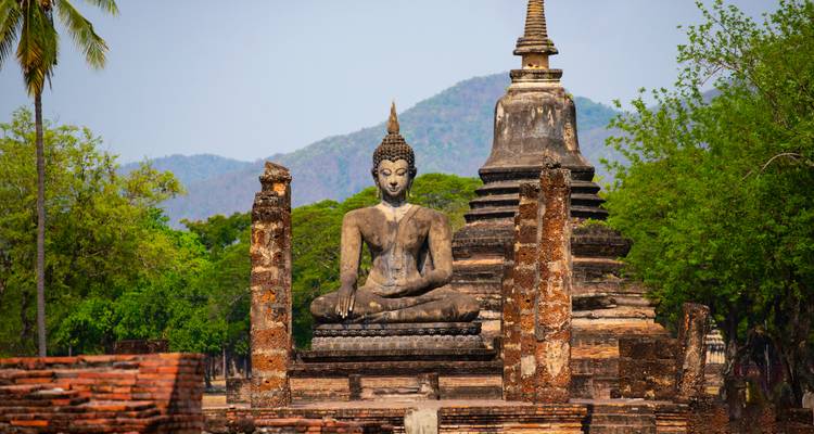 Statue de Bouddha assis ancien et ruines de grand chedi encadrées par des arbres verts avec des montagnes en arrière-plan.