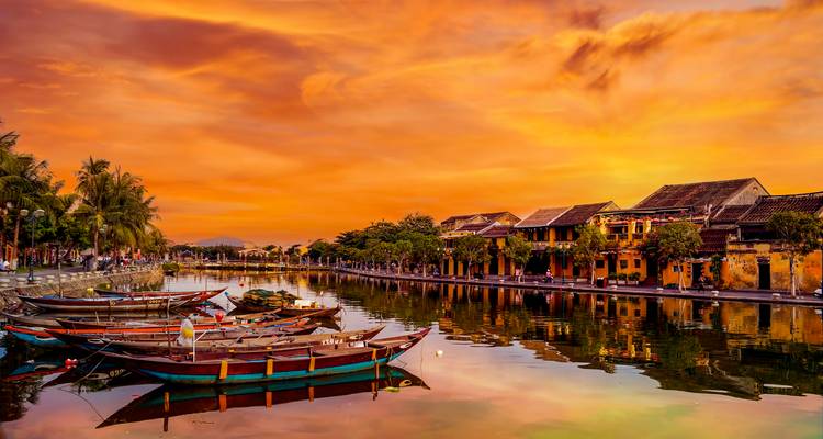 Las casas comerciales junto al río de Hoi An brillan bajo un cielo al atardecer de color naranja dramático que se refleja en el agua.