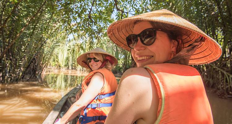 Dos mujeres que usan sombreros cónicos y chalecos salvavidas sonríen mientras navegan por canales de la selva llenos de lodo.